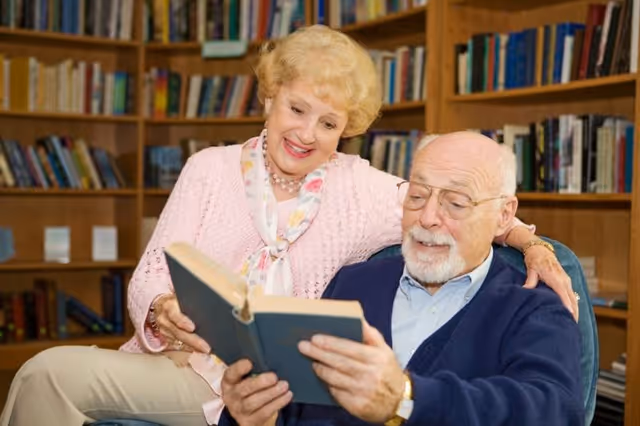 An elderly couple sitting together in a cozy library or study room. The man is seated in a chair holding and reading a book, while the woman stands beside him with her arm around his shoulder, smiling and looking at the book. Bookshelves filled with books are visible in the background.