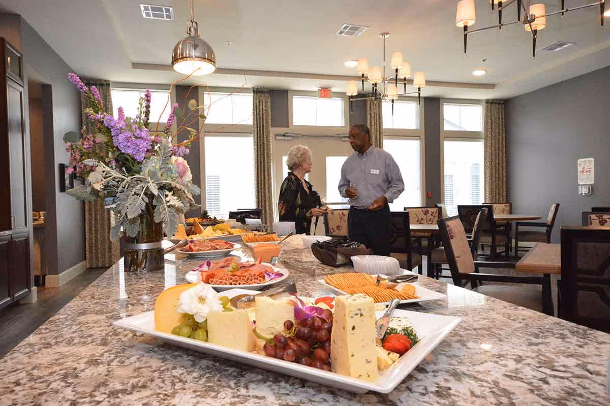 A dining area in The Crossings Retirement Community featuring a granite countertop with a platter of assorted cheeses, grapes, and crackers. In the background, two people are conversing near tables and chairs with floral upholstery. The room has large windows with curtains and modern light fixtures.