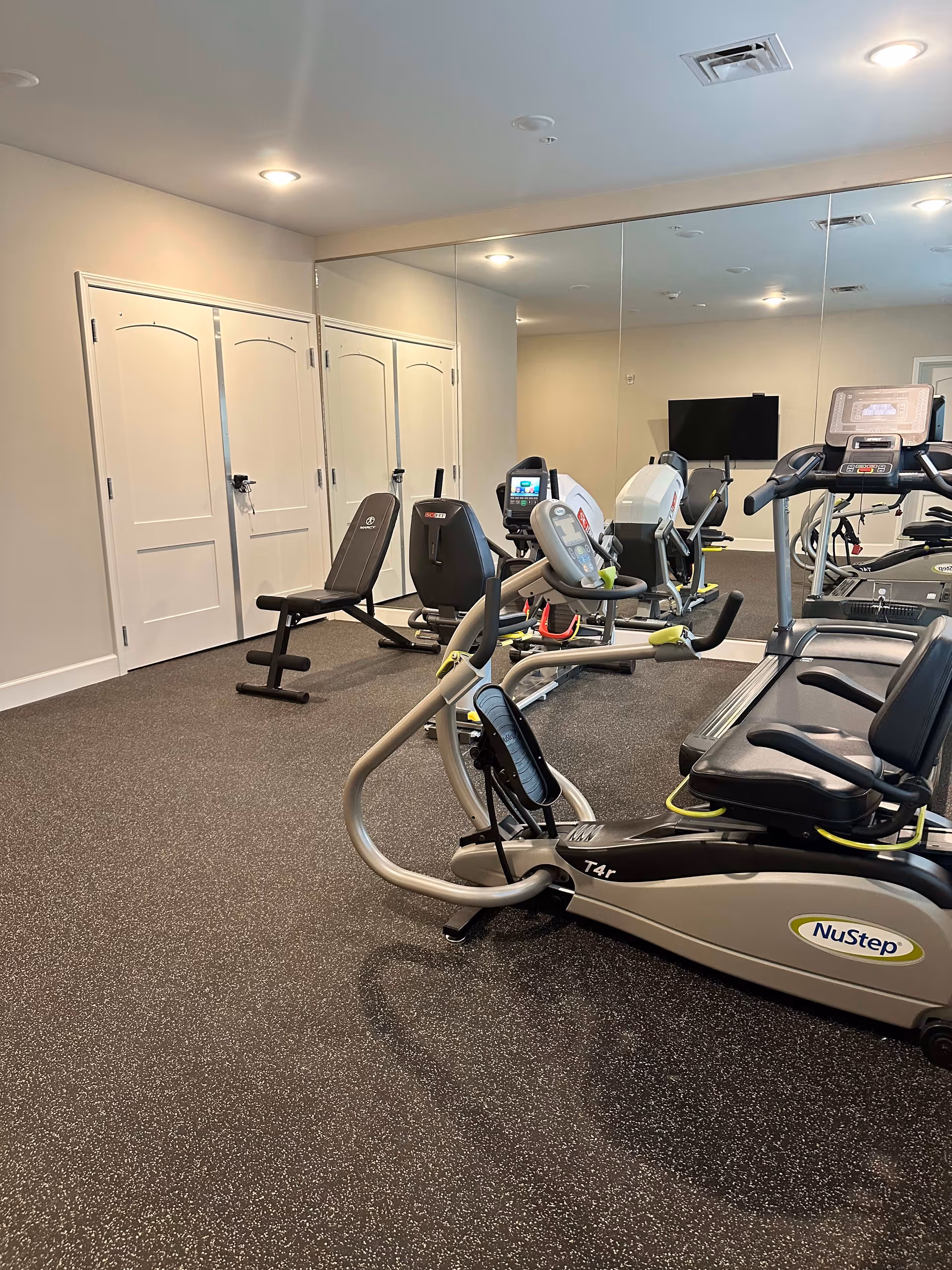 Interior view of a fitness room with exercise equipment including a NuStep recumbent cross trainer, a treadmill, and a weight bench. The room has a large wall mirror reflecting the equipment and a TV mounted on the wall. The floor is covered with speckled rubber gym flooring and the walls are painted light beige.