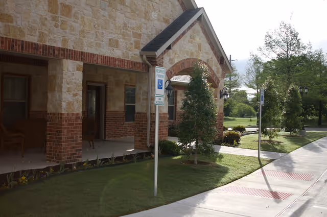 Front exterior of a stone-and-brick senior living building with a covered porch, accessible parking signs, landscaped lawn, and sidewalk.