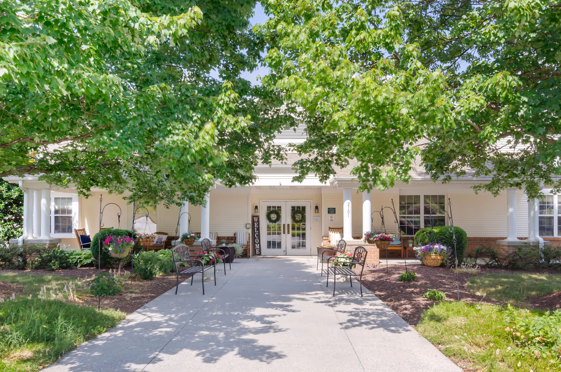 Front entrance of a senior living facility with a concrete walkway leading to double glass doors. The entrance is shaded by large green trees and flanked by white columns. There are benches and hanging flower baskets on either side of the walkway, with a welcome sign near the door.