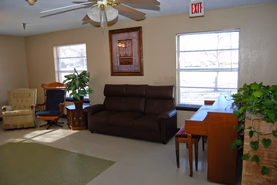 A cozy living room area with a brown leather sofa, two armchairs, a wooden side table with a potted plant, a piano with a bench, and two large windows letting in natural light. A ceiling fan with lights is mounted on the ceiling, and a framed artwork hangs on the wall between the windows. There is an exit sign above one window and a leafy green plant on a brick surface near the piano.