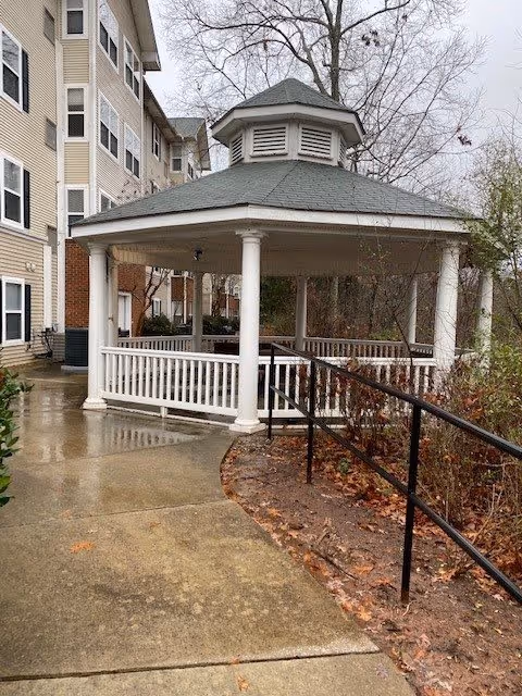A white gazebo with a gray roof situated on a wet concrete pathway next to a multi-story beige building. The gazebo has white railings and columns, with a black handrail along the pathway. Leafless trees and some bushes are visible in the background under an overcast sky.