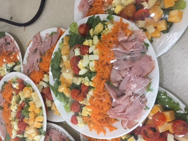 Several plates of salad covered with plastic wrap, each containing lettuce, cherry tomatoes, shredded carrots, diced cheese, and slices of ham, arranged on a countertop.