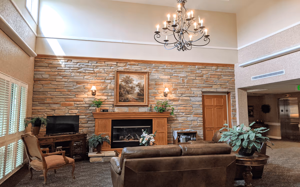 A cozy living room area in a senior living facility with a stone fireplace, a wooden mantel decorated with plants and a framed painting above it. There is a chandelier hanging from the high ceiling, a leather sofa facing the fireplace, an armchair near a window with white shutters, a TV on a wooden stand, and a round wooden table with a potted plant. The room has warm lighting and a carpeted floor.