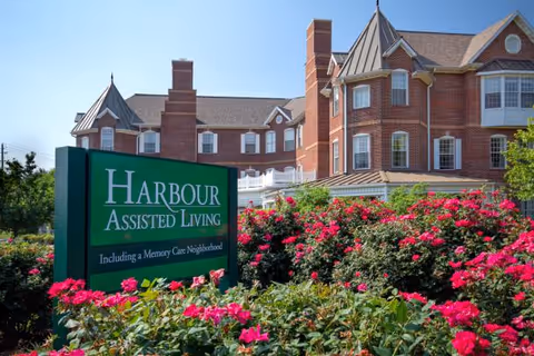 Exterior view of Harbour Assisted Living facility with a large green sign in front surrounded by blooming pink flowers and a multi-story brick building in the background under a clear blue sky.