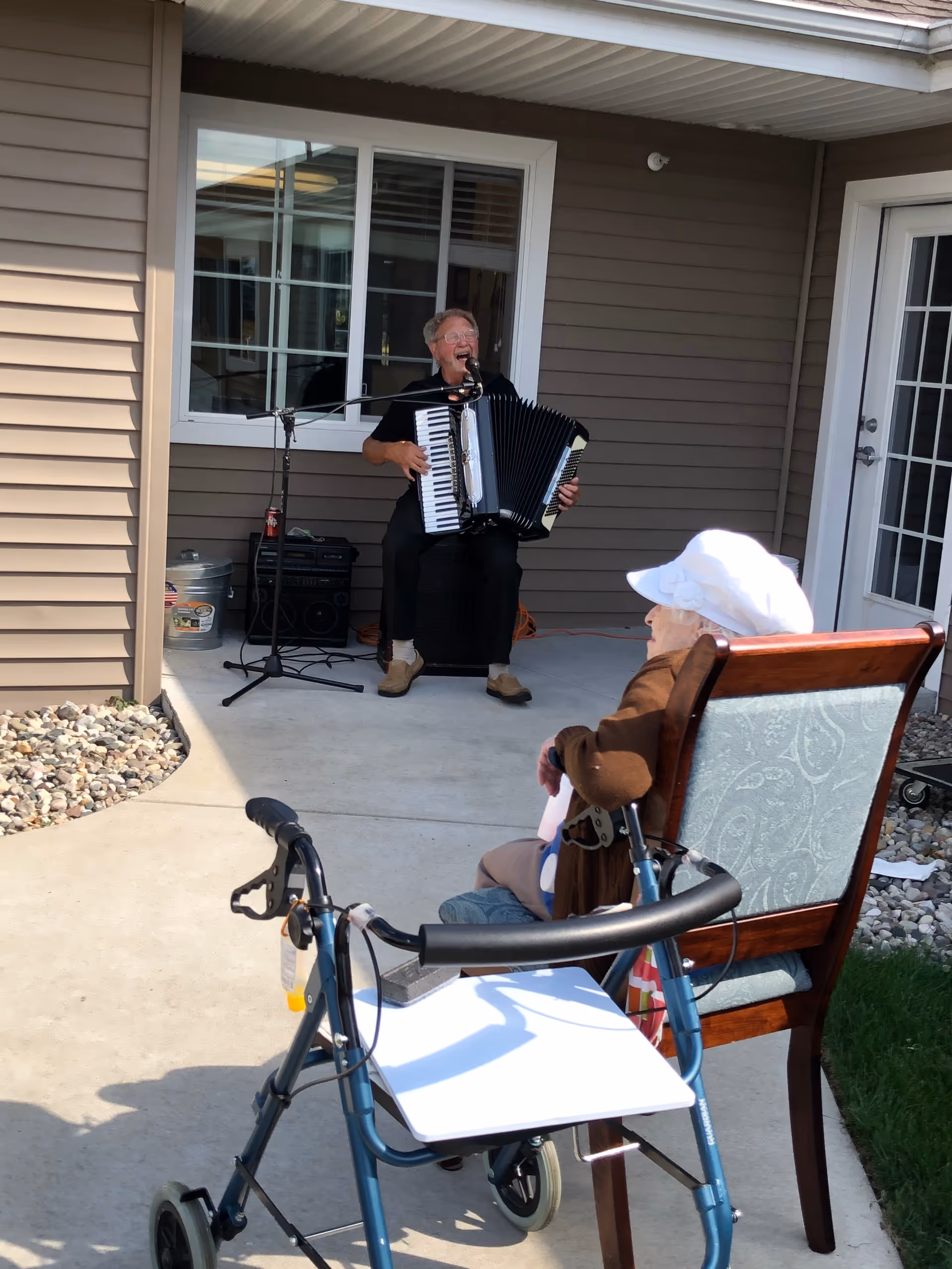 An elderly woman wearing a white hat sits in a chair outside on a patio, watching a man playing an accordion and singing into a microphone. The man is seated near a window and door of a building with beige siding. A walker is positioned near the woman.