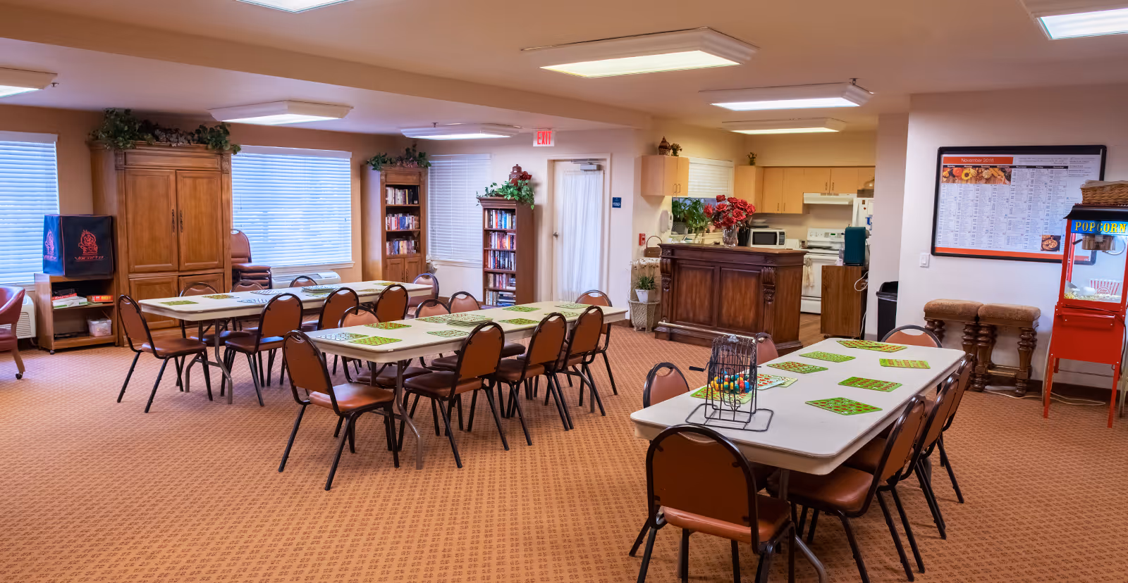 A spacious common area with several tables and chairs arranged for group activities or dining. The tables have green bingo cards and a bingo cage with colorful balls. The room has carpeted floors, large windows with blinds, bookshelves filled with books, a popcorn machine, and a kitchen area with wooden cabinets and appliances in the background. The walls are decorated with plants and a large calendar.