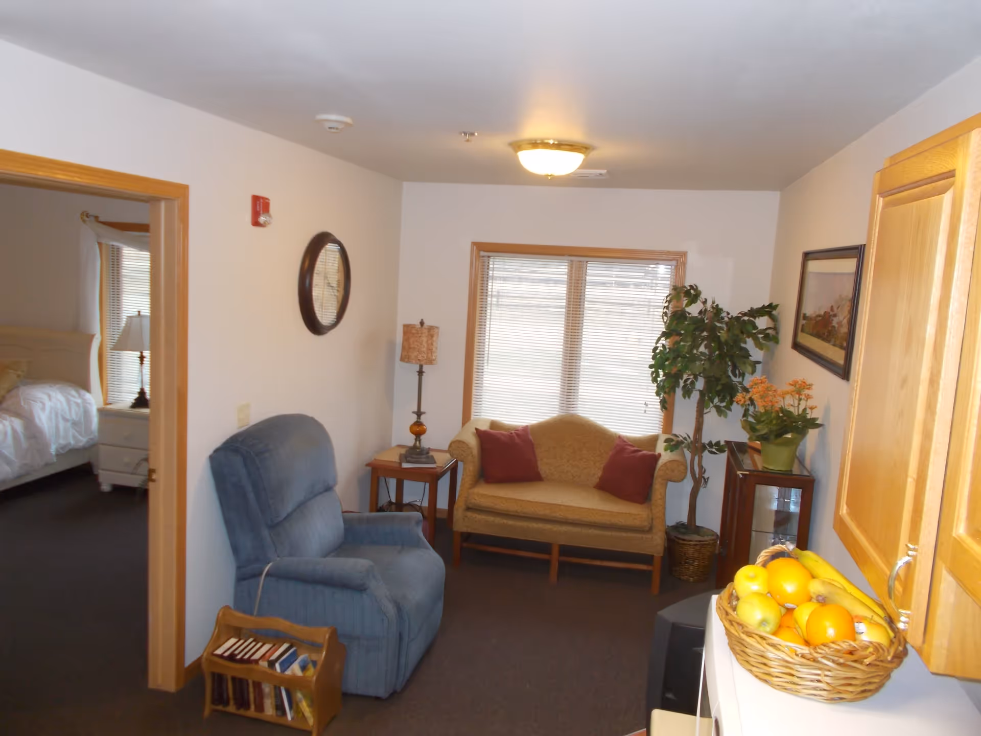Cozy living room with a blue recliner, a loveseat with pillows under a window, side tables and plants, and a basket of fruit on a nearby counter with a bedroom visible through a doorway.