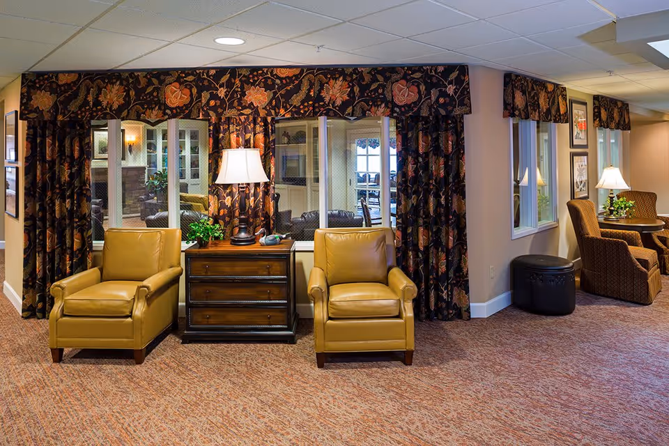 A cozy seating area in a memory care community featuring two tan leather armchairs separated by a wooden side table with a lamp and a small plant. Behind the seating area are large windows with floral-patterned curtains. To the right, there is another seating area with two upholstered chairs and a round table with a lamp and plant. The carpet is a warm, textured pattern and the walls are painted beige.