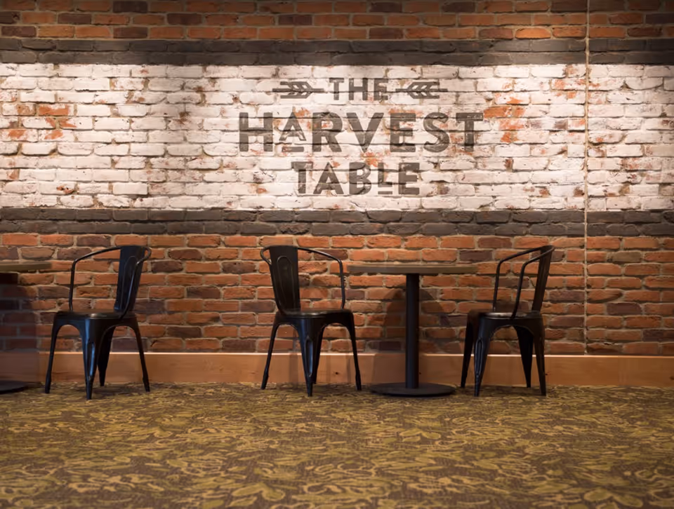A dining area with a brick wall background featuring the words 'The Harvest Table' painted on a whitewashed section of the wall. In front of the wall are three black metal chairs and a small round table on a patterned carpeted floor.