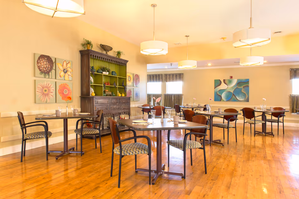 A bright dining room with wooden floors, several tables each set with plates, glasses, and flowers in small vases. The room features multiple hanging light fixtures, floral artwork on the walls, and a large wooden cabinet with decorative items.