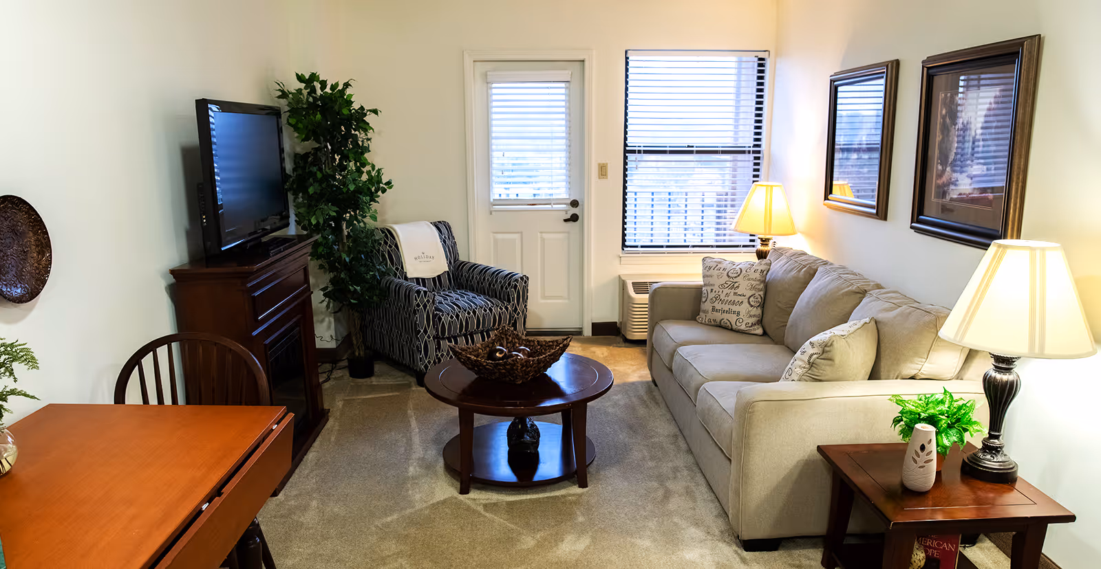 Small furnished living room with a beige sofa, patterned armchair, TV on a stand, coffee table, and lamps.
