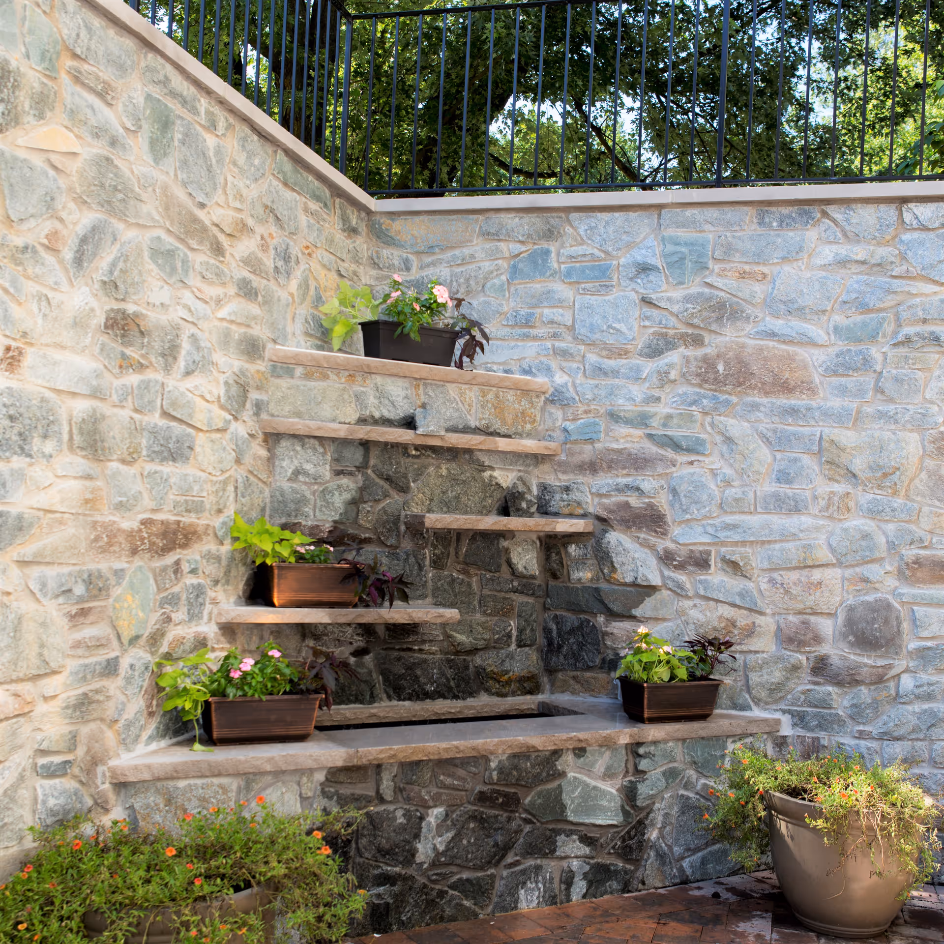 Stone wall with built-in tiered shelves holding potted plants, surrounded by additional potted plants on the ground, with a black metal fence and trees visible above the wall.