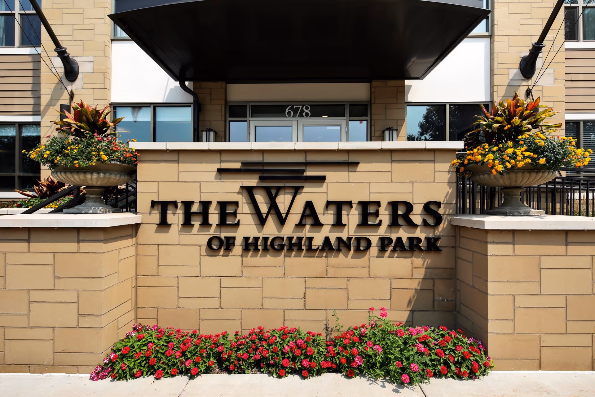Entrance sign for The Waters of Highland Park facility mounted on a beige brick wall with flower beds and large planters with colorful flowers on either side. The building entrance with glass doors and the number 678 is visible in the background.