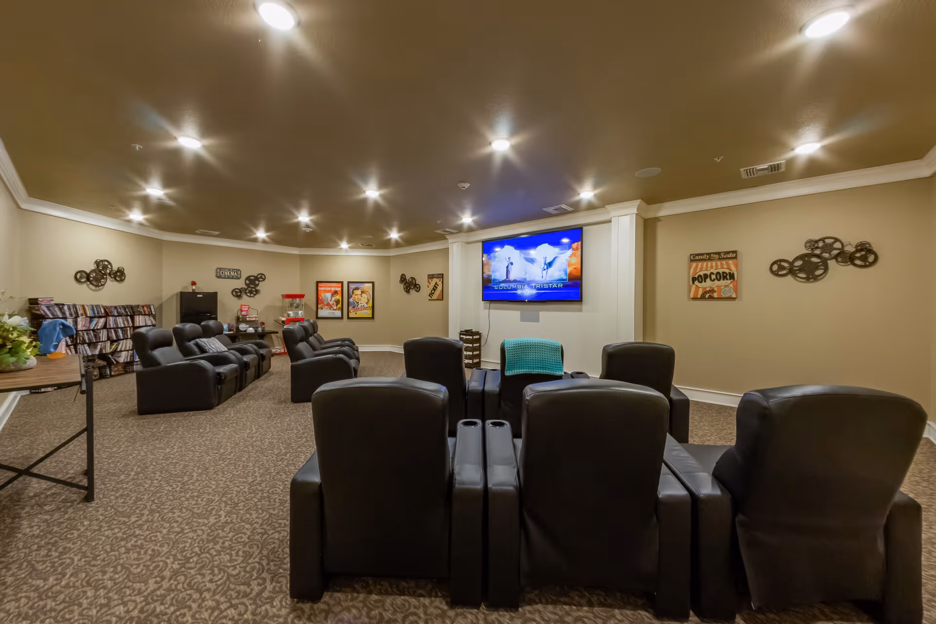 A cozy communal media room with rows of black recliner seats facing a wall-mounted TV and popcorn machine.