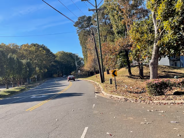 A curved road lined with trees on both sides under a clear blue sky. There is a car driving away in the distance, utility poles with power lines along the right side, and a yellow road sign indicating a curve. The area appears to be suburban with some greenery and a small hill on the right.