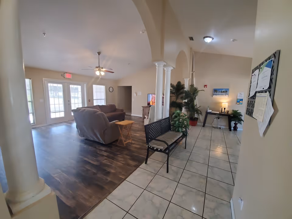 Interior view of a senior living facility lounge area with beige walls and a mix of tiled and wooden flooring. The space features a cushioned bench, two sofas, a small wooden side table, a ceiling fan with lights, potted plants, a TV on a wooden stand, and a bulletin board with papers on the wall. There is natural light coming through glass doors and windows.