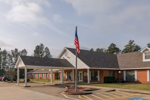 Exterior view of a single-story brick healthcare and rehabilitation center building with a covered entrance and an American flag on a flagpole in front. There are trees in the background and a parking area in the foreground.
