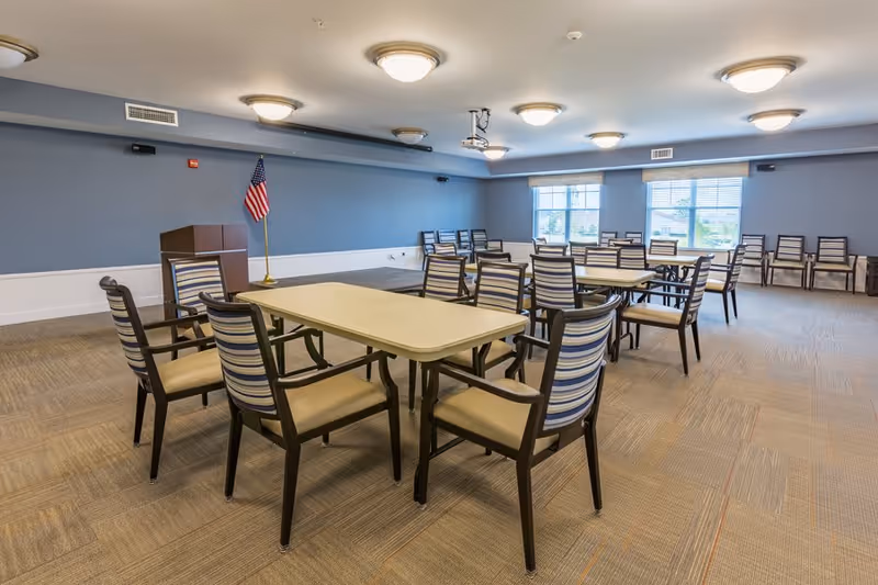 A spacious meeting or activity room with several tables and chairs arranged in rows. The walls are painted blue with white trim, and there is an American flag on a stand near a podium. Large windows allow natural light to enter the room, and ceiling lights provide additional illumination.