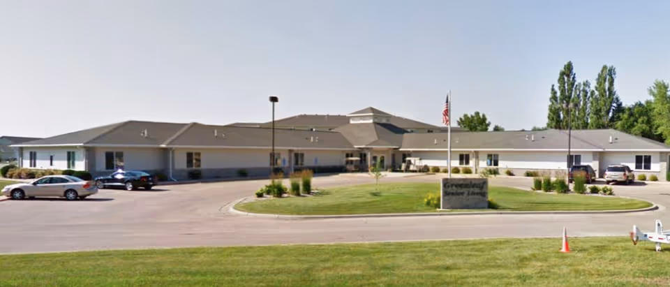 Single-story beige senior living facility with a circular driveway, flagpole, sign, and several parked cars.