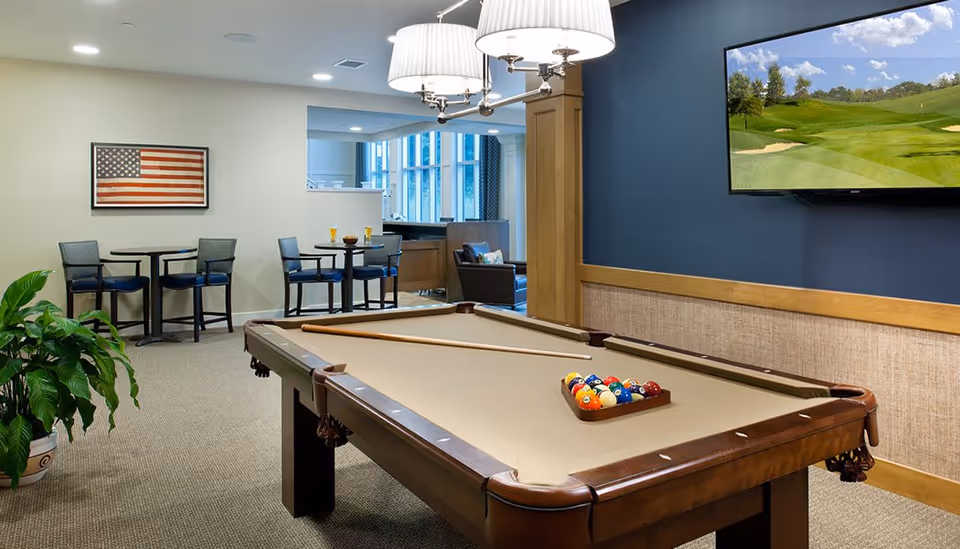 A recreational room featuring a pool table with a full set of billiard balls and a cue stick on top. The room has a blue accent wall with a mounted flat-screen TV displaying a golf course. There are two small round tables with chairs near a wall decorated with a framed American flag. A large green potted plant is visible on the left side, and a comfortable seating area with armchairs is seen in the background.