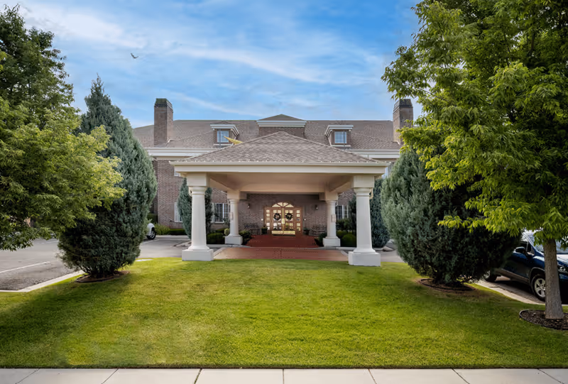 Front entrance of a brick senior living building featuring a covered porte-cochère with columns, flanked by trees and a manicured lawn.