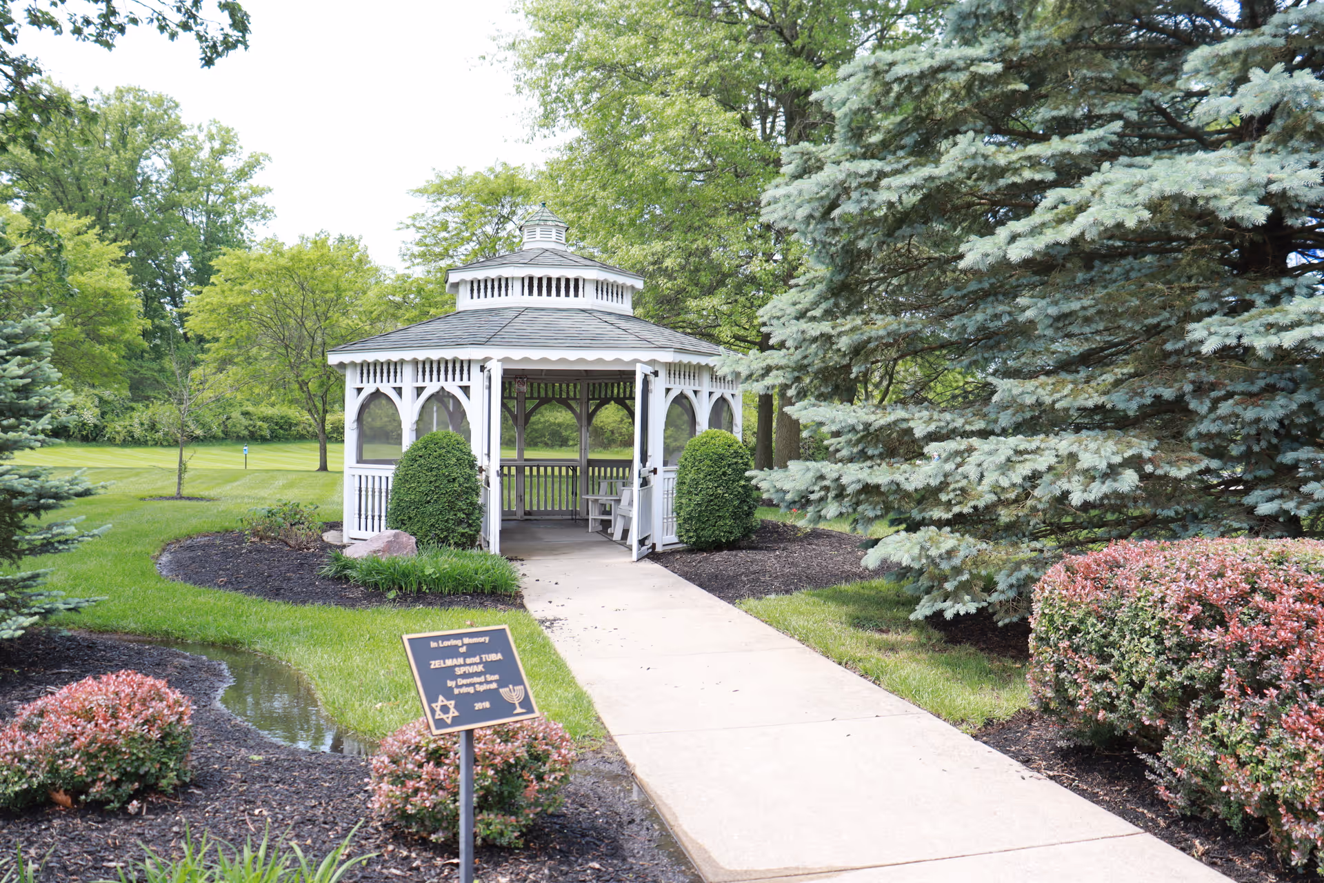 A white wooden gazebo with a shingled roof is situated at the end of a paved walkway in a landscaped garden area. The surrounding greenery includes trimmed bushes, trees, and well-maintained grass. A memorial plaque is visible near the walkway in front of the gazebo.