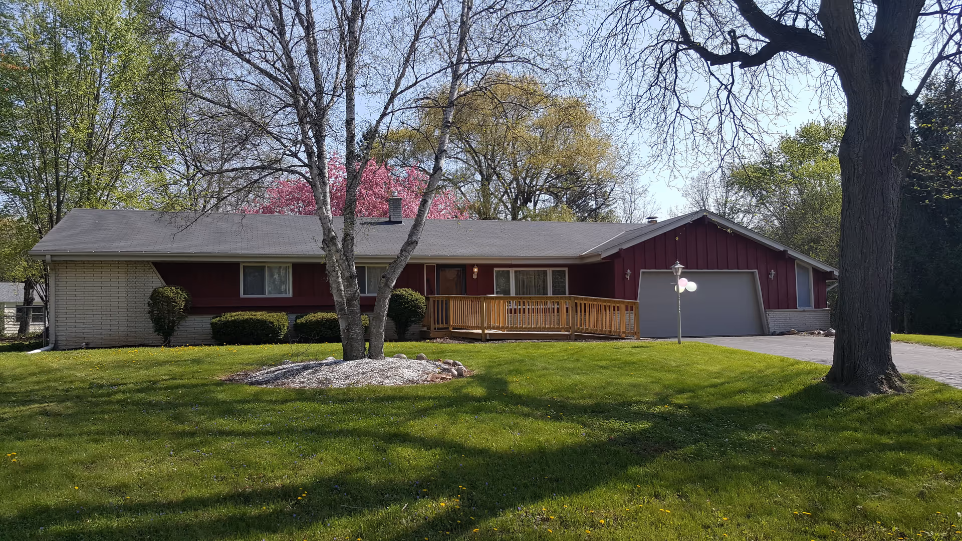 Single-story residential building with a gray roof and red siding, surrounded by green grass and trees with some pink blossoms. There is a wooden ramp leading to the front door and a driveway with a garage on the right side.