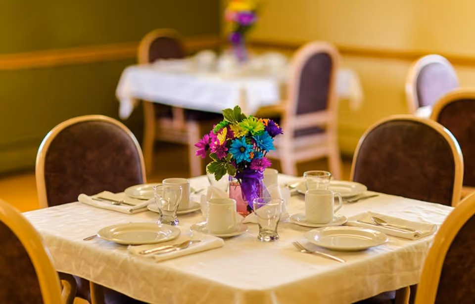 A dining table set for four with white plates, cups, glasses, and silverware on a white tablecloth. A colorful bouquet of flowers in a purple vase is placed in the center of the table. In the background, another similarly set table is visible with chairs around it.