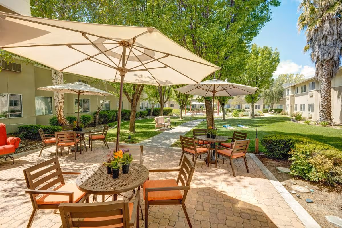 Outdoor patio area at Creston Village with several round tables and chairs under large beige umbrellas. The patio is paved with bricks and surrounded by green grass, trees, and shrubs. Residential buildings are visible in the background under a clear blue sky.