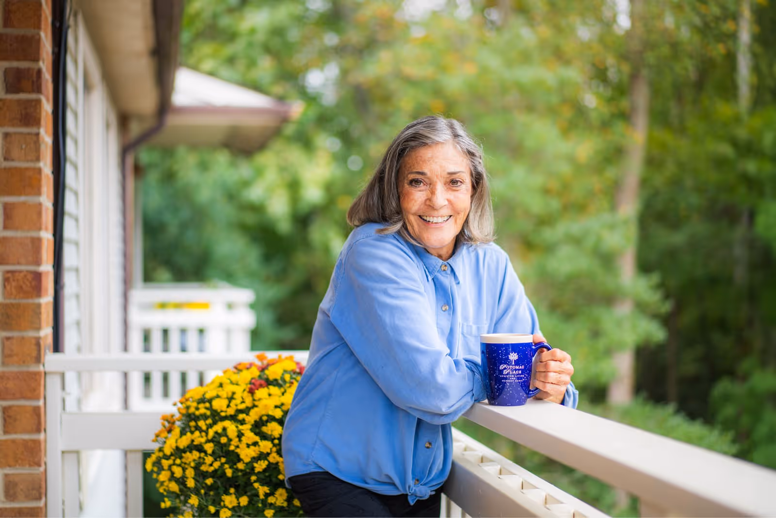 An elderly woman with gray hair wearing a blue shirt is smiling and leaning on a white balcony railing outdoors. She is holding a blue mug with the text 'Potomac Place Assisted Living and Memory Care'. Behind her, there is a pot of yellow flowers and green trees in the background.