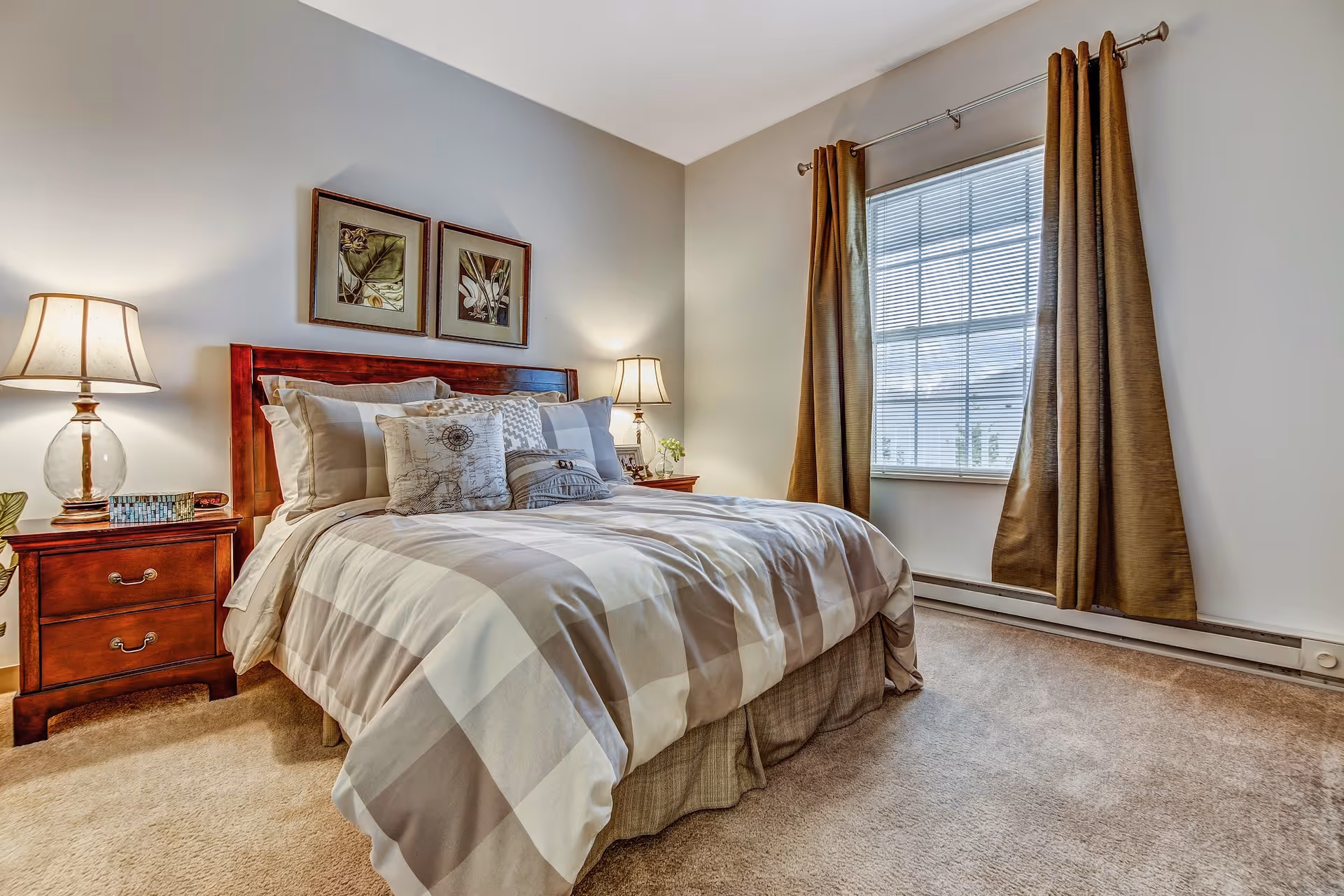 A cozy bedroom with a neatly made bed featuring a checkered comforter and multiple pillows. There are two wooden nightstands on either side of the bed, each with a lamp. Above the bed, two framed botanical prints hang on the wall. A window with brown curtains allows natural light into the room, which has beige carpeting and light-colored walls.