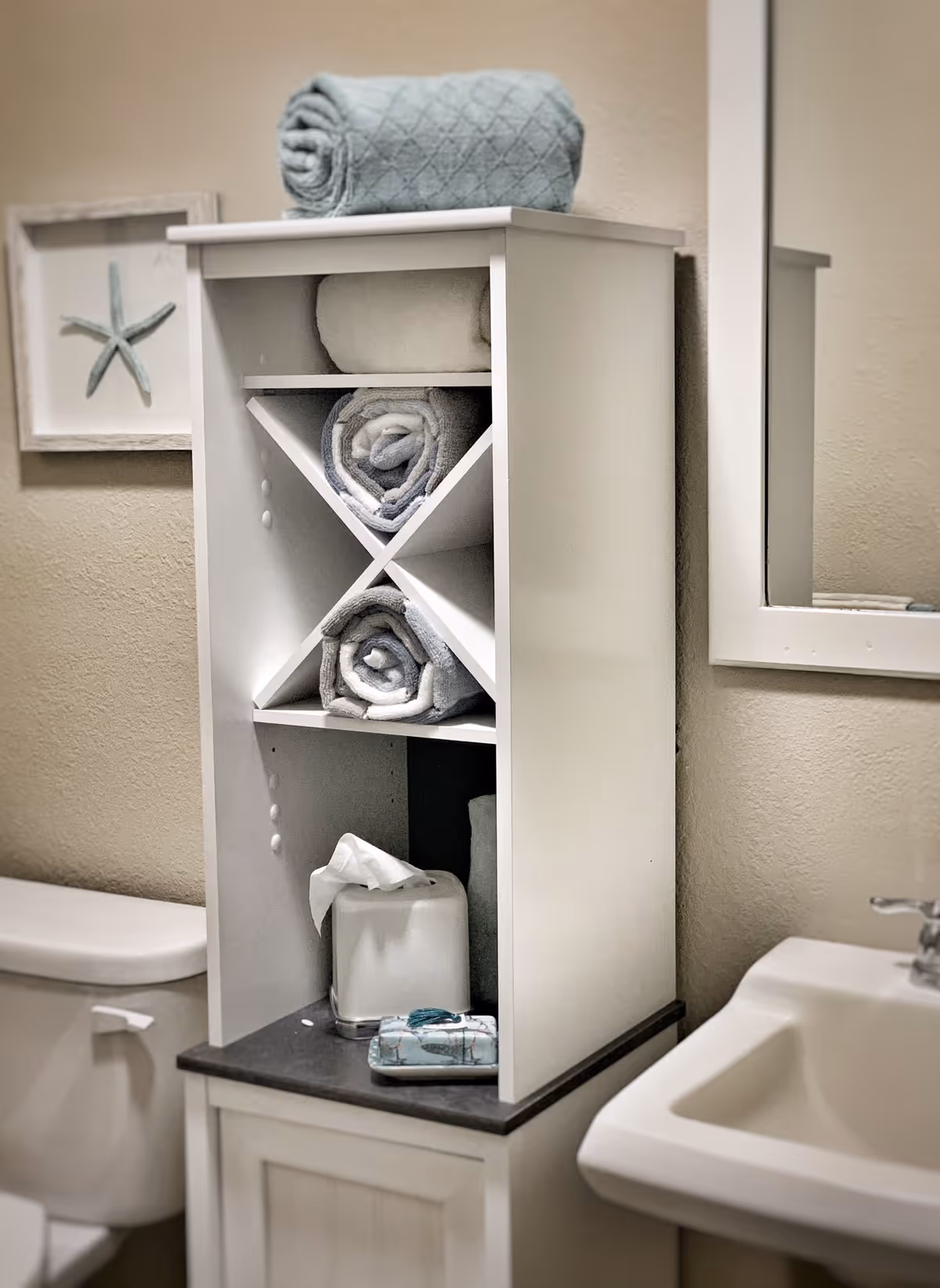 White shelving cabinet above a toilet holding rolled towels and a tissue box beside a sink and mirror.