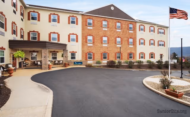 Exterior view of a three-story senior living facility building with a covered entrance, multiple windows with red shutters, an American flag on a flagpole, and a paved driveway with landscaped areas.