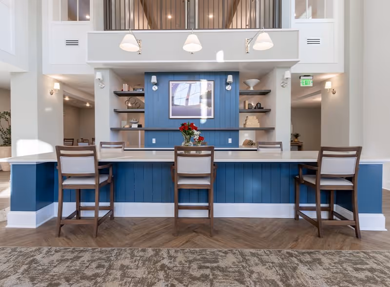 A modern reception or common area with a blue paneled counter and three wooden chairs with light cushions. Behind the counter are built-in shelves with decorative items and a framed picture on a blue accent wall. The space has a high ceiling with hanging lights and a carpeted floor in the foreground.