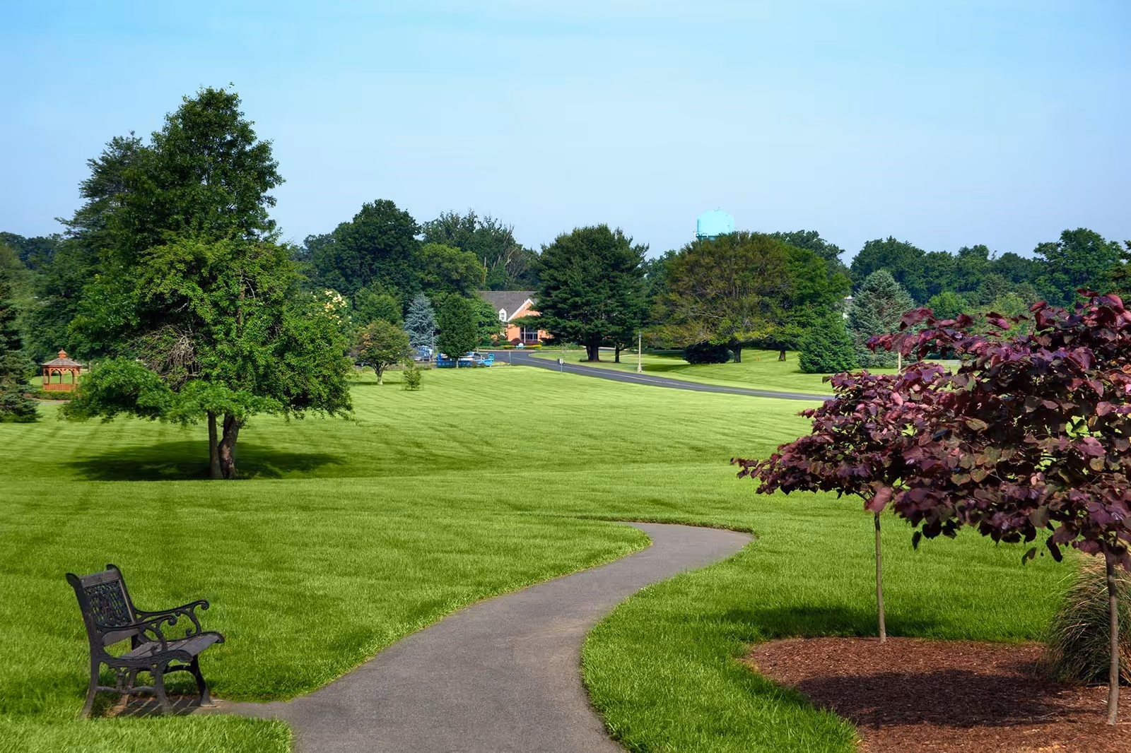 A winding paved pathway through a well-maintained green lawn with various trees and a bench on the left side. In the background, there are more trees, a small gazebo, and a building partially visible under a clear blue sky.