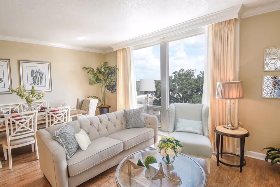A bright and cozy living room with a beige tufted sofa adorned with decorative pillows, a light blue armchair with a pillow, and a round glass coffee table with floral and geometric decorations. Behind the sofa is a dining area with a wooden table, white chairs, and floral artwork on the wall. Large windows with beige curtains let in natural light and offer a view of trees outside.