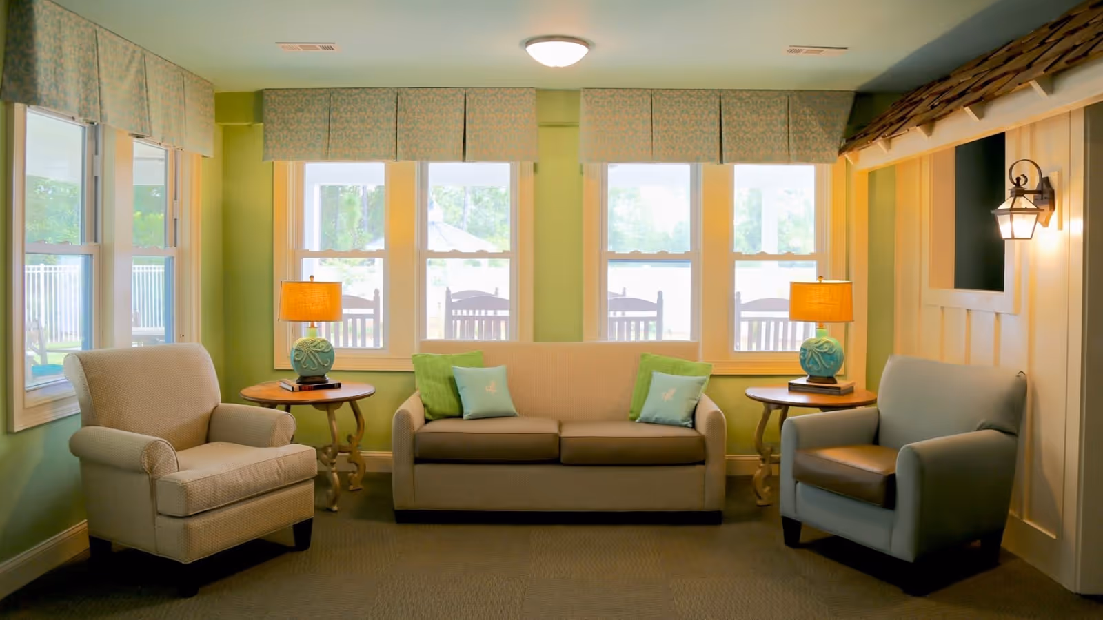 A cozy sitting area in a senior care facility with a beige sofa adorned with green and light blue pillows, flanked by two armchairs in beige and light blue. Two round wooden side tables each hold a lamp with a blue base and an orange shade. The room has light green walls and three large windows with patterned valances, allowing natural light to brighten the space. A decorative wall sconce is mounted on the right wall.
