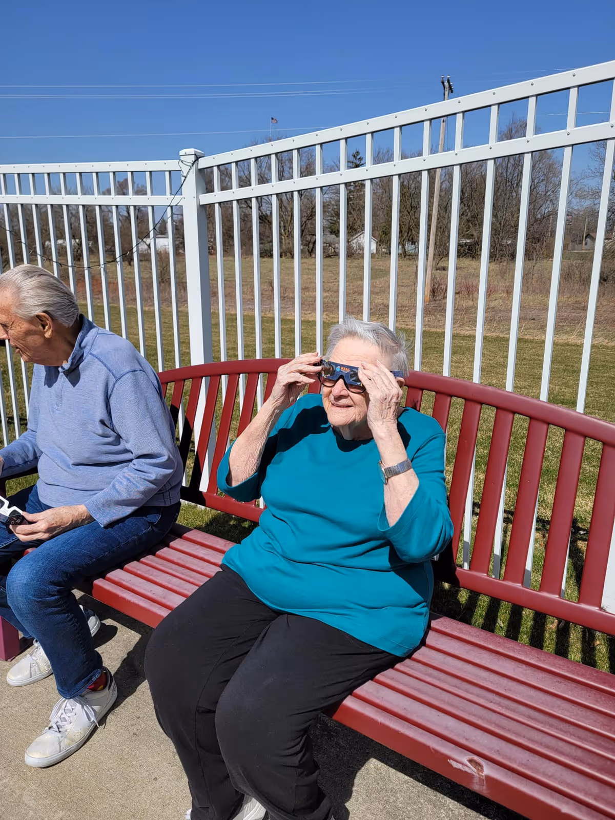 Two elderly individuals sitting on a red bench outdoors near a white metal fence. One person is wearing a teal top and black pants, holding solar eclipse glasses up to their eyes. The other person is wearing a light blue long-sleeve shirt and jeans, looking down at something in their hands. The background shows a grassy area and trees under a clear blue sky.