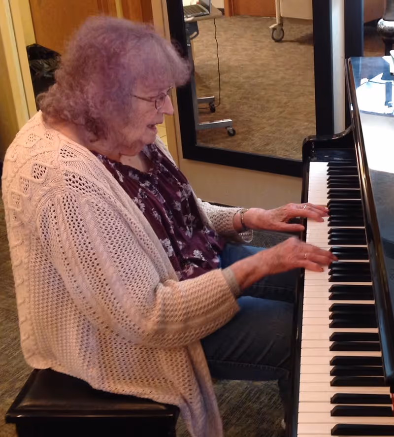 An elderly woman with curly gray hair and glasses is sitting on a bench playing a piano indoors. She is wearing a light pink knitted cardigan over a floral blouse and blue jeans. A large mirror and a door are visible in the background.