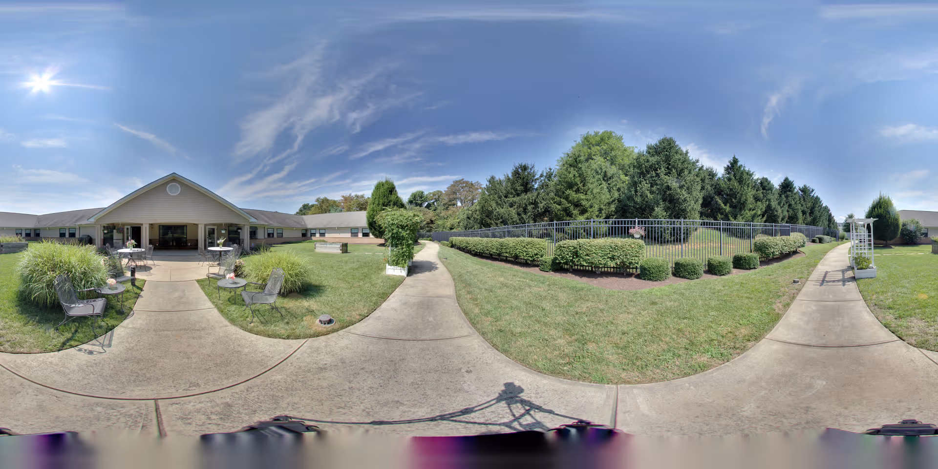 Outdoor courtyard of a senior living facility with a central concrete walkway leading to a low building entrance, patio tables and chairs, manicured hedges and trees under a blue sky.