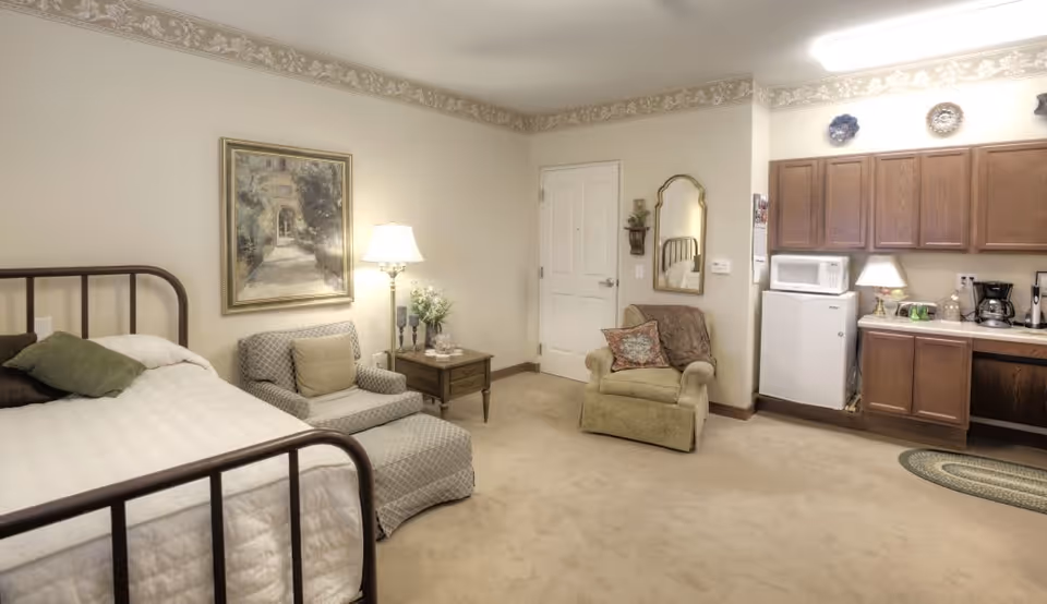 A cozy room featuring a bed with a metal frame, a cushioned armchair with a matching ottoman, a side table with a lamp and decorative items, and a kitchenette area with wooden cabinets, a mini refrigerator, microwave, coffee maker, and a small rug on the carpeted floor. The walls are light-colored with a decorative border near the ceiling, and a framed painting hangs above the armchair.