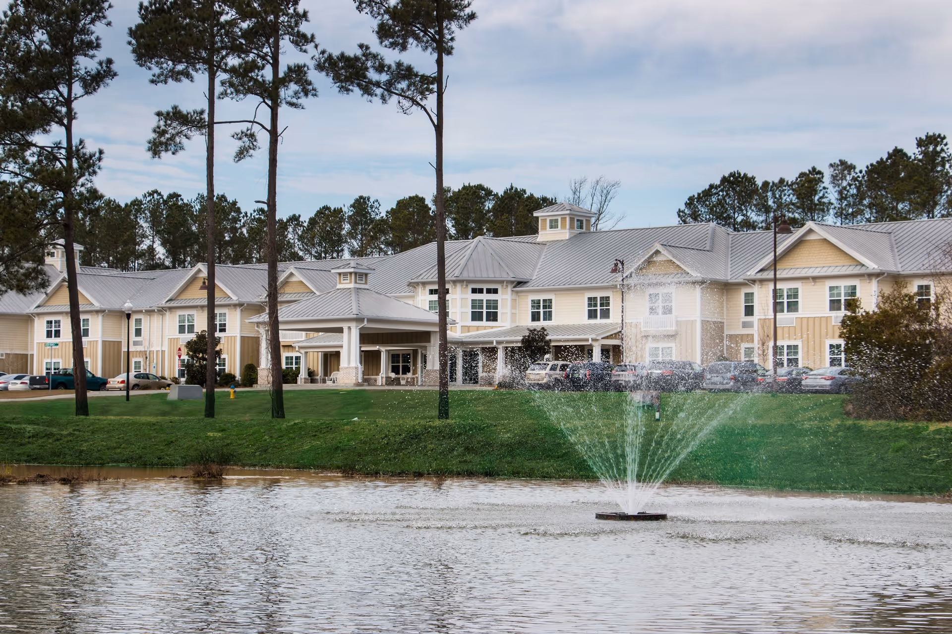 View of Arbor Landing at Ocean Isle senior living facility with a large pond and a water fountain in the foreground, surrounded by green grass and tall pine trees, under a partly cloudy sky.