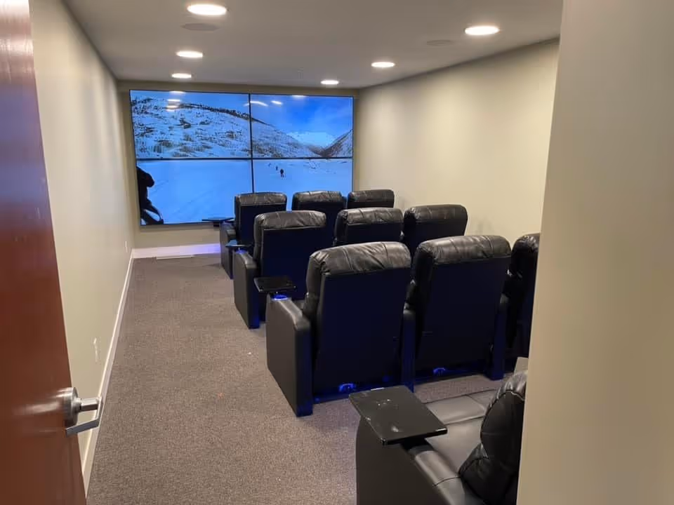 A small theater room with two rows of black leather recliner chairs facing a large multi-panel screen displaying a snowy mountain scene. The room has beige walls, recessed ceiling lights, and a carpeted floor.