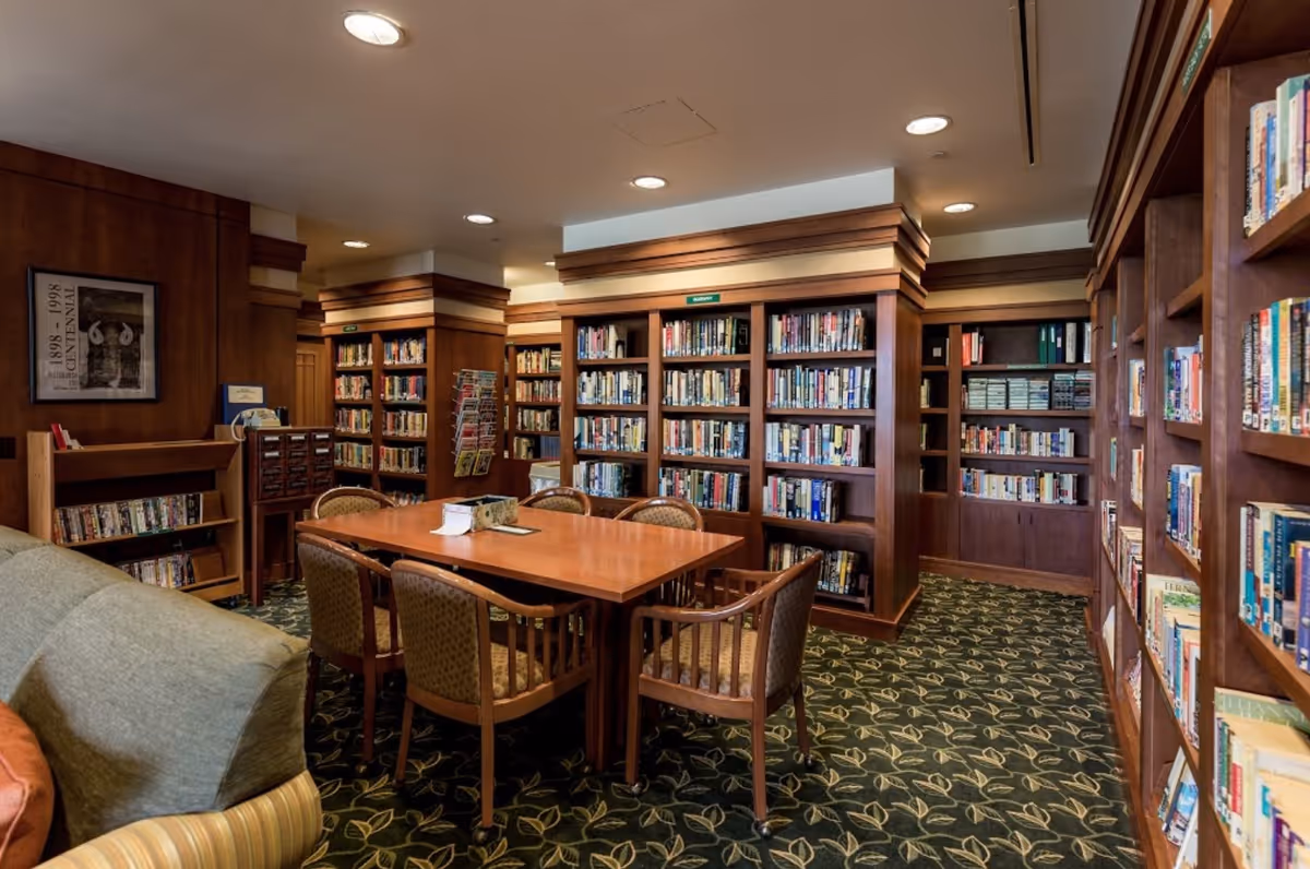 A cozy library room with wooden bookshelves filled with books lining the walls. In the center, there is a wooden table surrounded by six cushioned chairs. The room has a patterned carpet with leaf designs and recessed ceiling lights providing illumination. A framed picture hangs on the left wall above a small bookshelf.