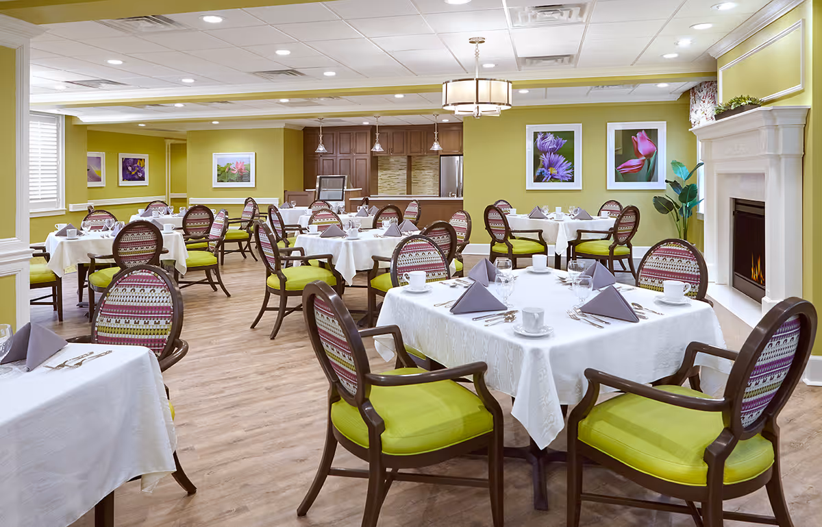 A bright and elegant dining room with multiple round tables covered in white tablecloths, set with cups, glasses, silverware, and folded gray napkins. The chairs have green cushions and patterned backs. The room has light green walls adorned with framed floral artwork, a fireplace on the right side, and a wooden cabinet area in the back. The floor is wooden, and the ceiling has recessed lighting and a hanging light fixture.