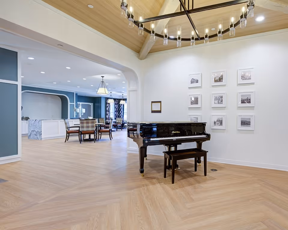 Interior view of a senior living facility featuring a black grand piano with a bench on a light wood floor. The walls are white with a series of framed black and white photographs arranged in a grid. In the background, there is a dining area with tables and chairs, blue walls, and pendant lighting fixtures hanging from the ceiling.