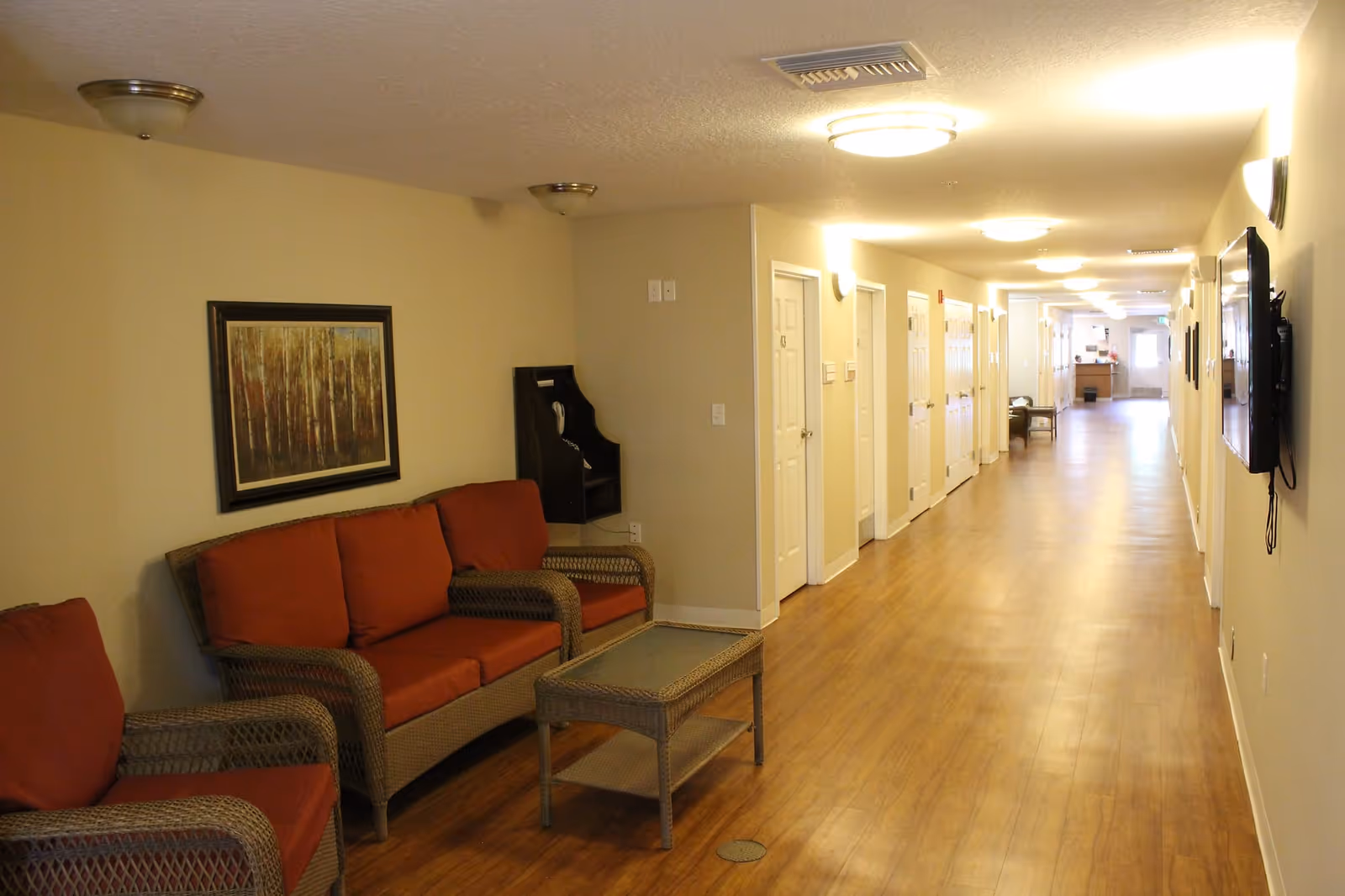 A long, well-lit hallway with wooden flooring and beige walls. On the left side, there is a seating area with wicker furniture including a sofa and two chairs with red cushions, and a glass-top coffee table. A framed painting hangs above the sofa. Several white doors line the hallway, and a wall-mounted telephone is visible near the seating area. A flat-screen TV is mounted on the right wall.