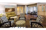 A cozy living room area in a senior care facility featuring four patterned armchairs arranged around a round ottoman, a green sofa against the wall, a landscape painting above the sofa, and large windows letting in natural light. The room has wooden flooring and double doors leading to another area.